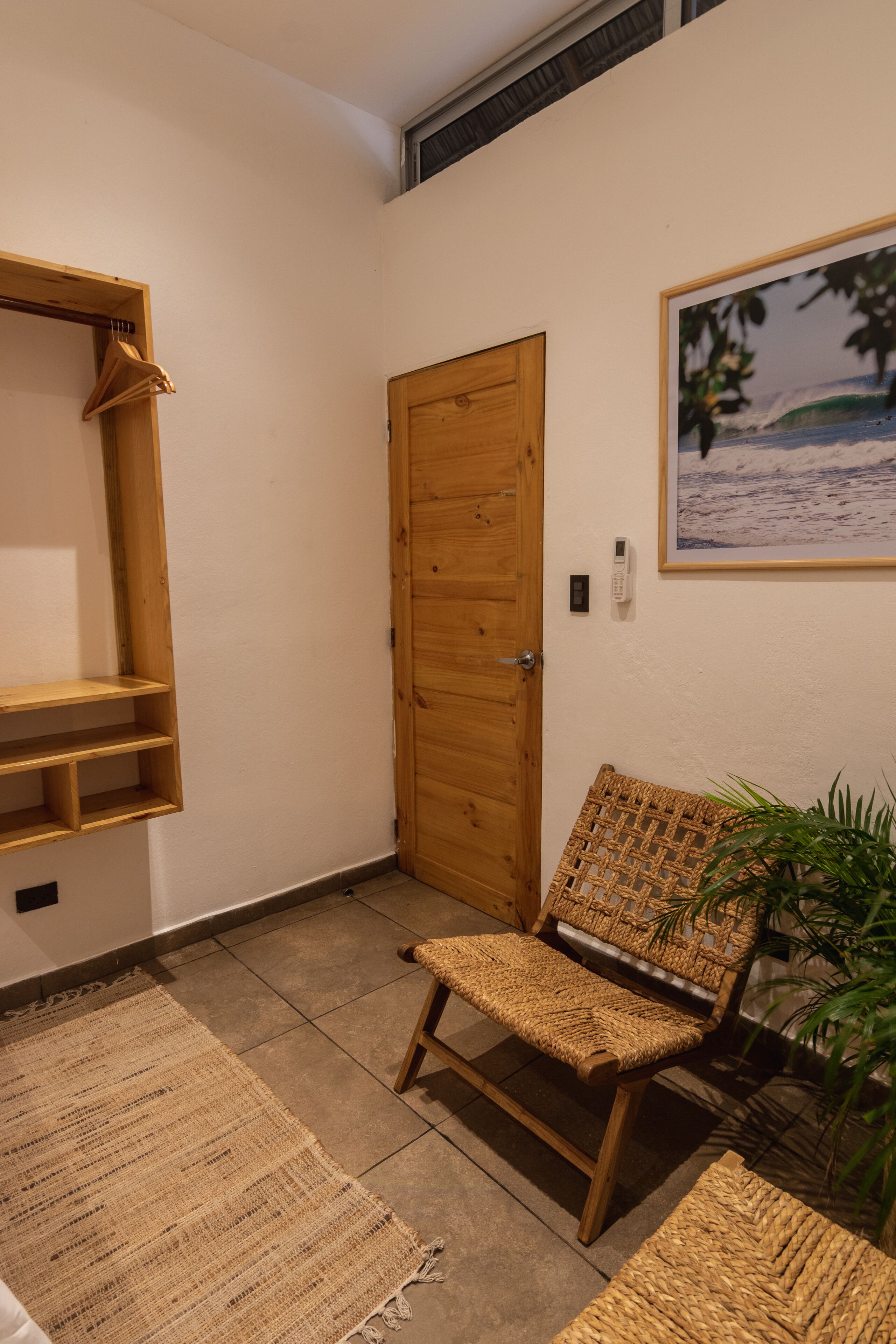 Una habitación con puerta de madera, un banco de mimbre, un estante pequeño y un cuadro enmarcado en la pared.
