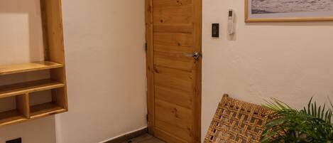 Una habitación con puerta de madera, un banco de mimbre, un estante pequeño y un cuadro enmarcado en la pared.