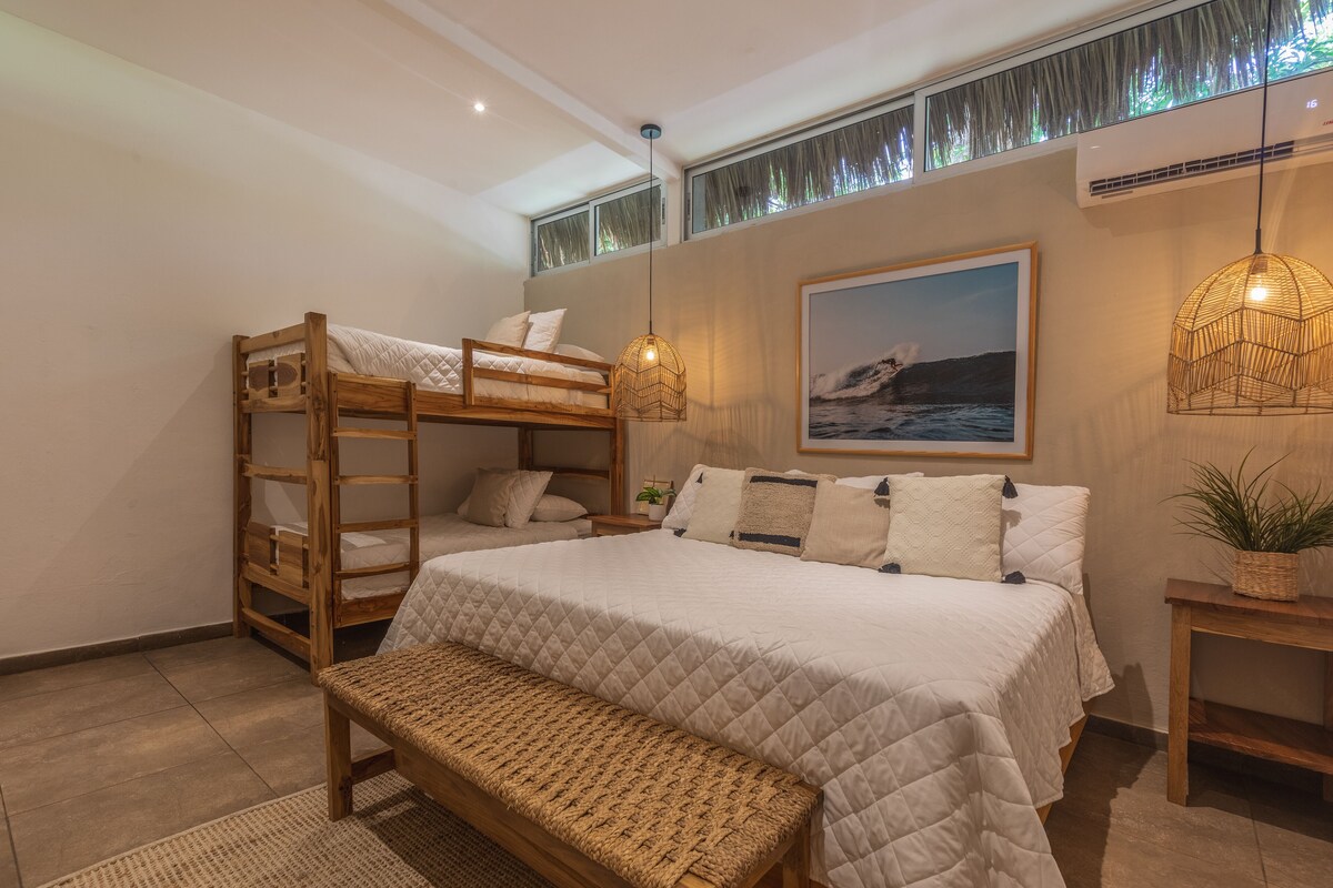 A bedroom with a bunk bed, a single bed, a bench, a hanging light fixture, a wall-mounted air conditioner, and a framed picture.