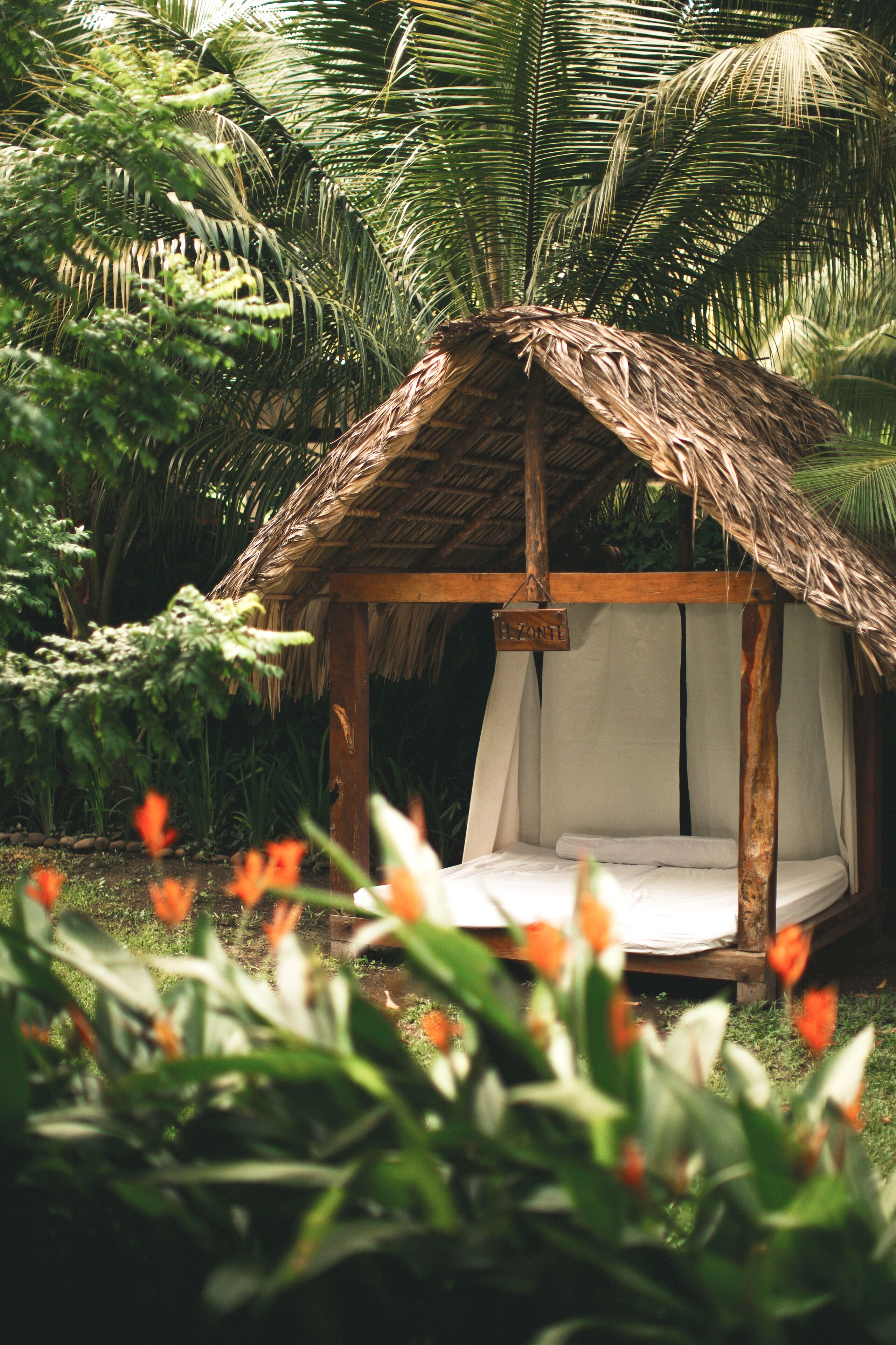 Uma cabana com telhado de palha e cama interior, rodeada por vegetação exuberante e plantas.