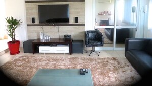Un salon avec un téléviseur à écran plat fixé au mur, une table basse, un canapé, un fauteuil et une plante en pot rouge.