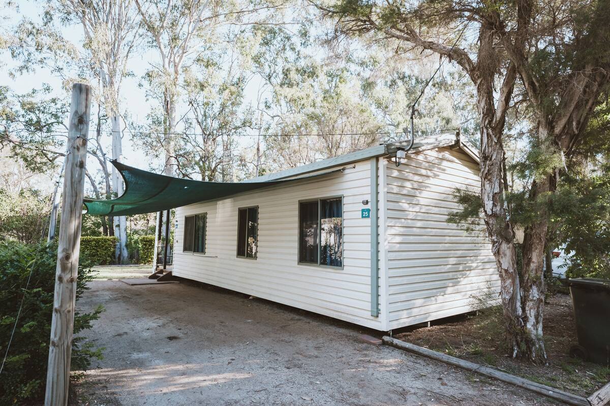 Una casa móvil de un piso, blanca, con toldo verde, rodeada de árboles y un camino de tierra.