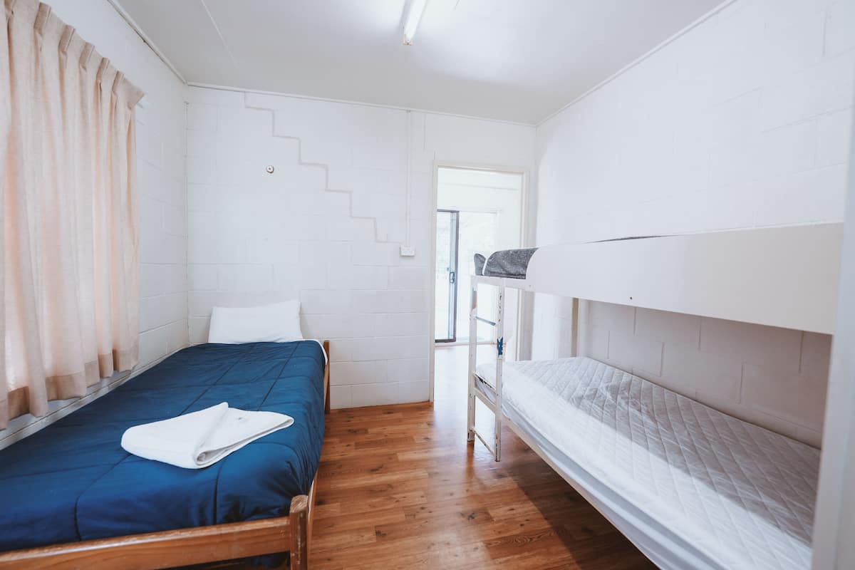 Una habitación con literas, una cama individual, suelo de madera, paredes blancas y una escalera que lleva a un espacio al aire libre.
