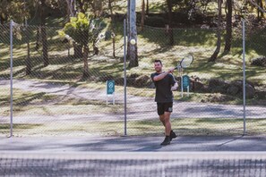 Ein Tennisspieler in einem Park mit einem Zaun und Bäumen im Hintergrund.