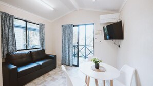 Una habitación moderna con un sofá negro, sillas blancas, una mesa redonda con una planta, un televisor montado en la pared y un balcón con barandilla.