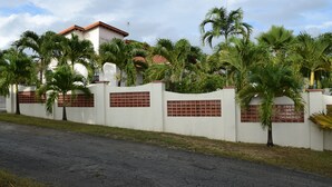 A white building with a red-tiled roof, surrounded by palm trees and a brick wall with decorative panels. - Eullen Villa (Arthurs Seat)