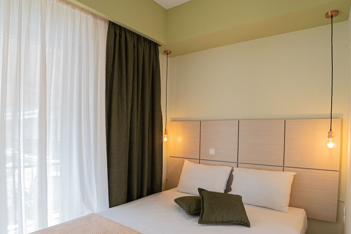 Uma cama bem arrumada com manta bege e almofadas, cabeceira com iluminação embutida e janela com cortinas brancas leves e uma cortina escura.