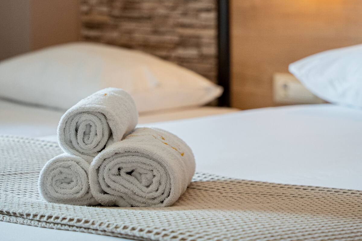 Cama bem arrumada com toalhas brancas enroladas, parede de pedra texturizada e luminária pendente.