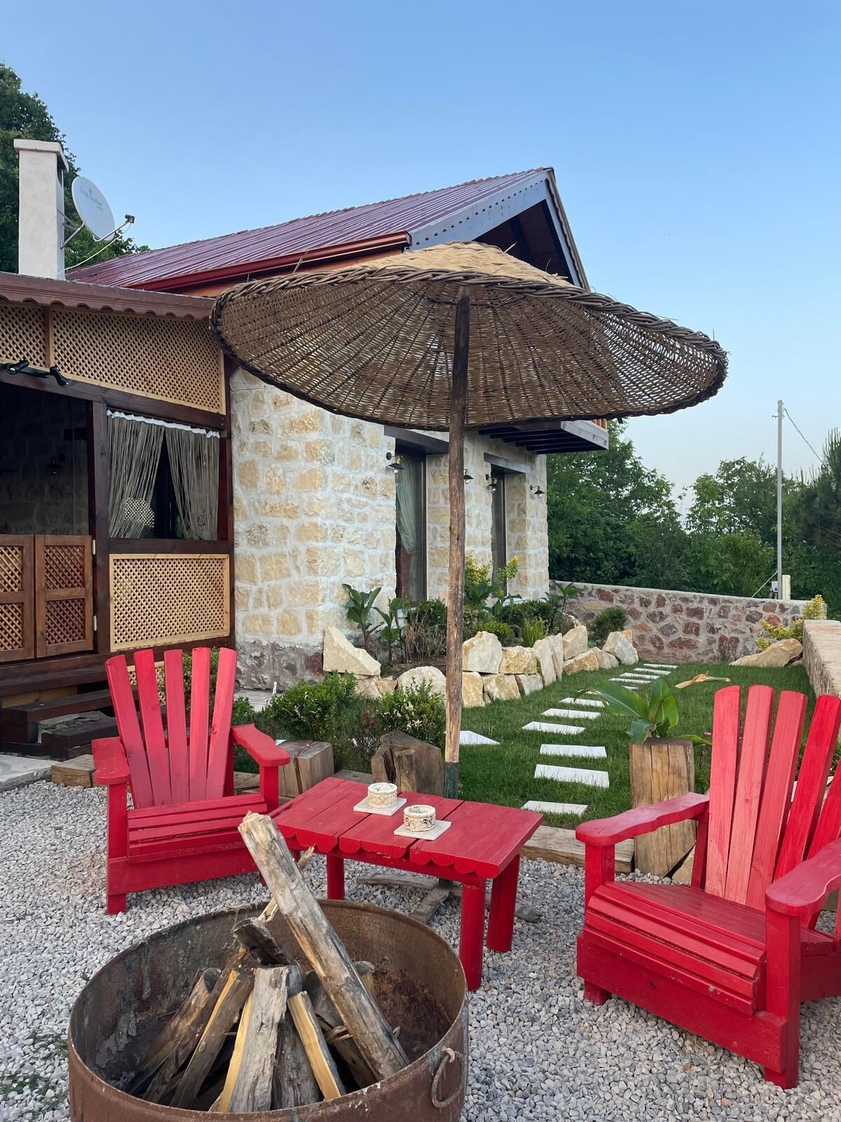 A stone building with a red roof, a thatched umbrella, red Adirondack chairs, a fire pit, and a stone pathway.