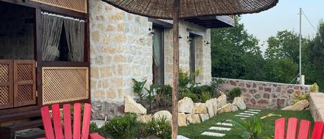 A stone building with a red roof, a thatched umbrella, red Adirondack chairs, a fire pit, and a stone pathway.