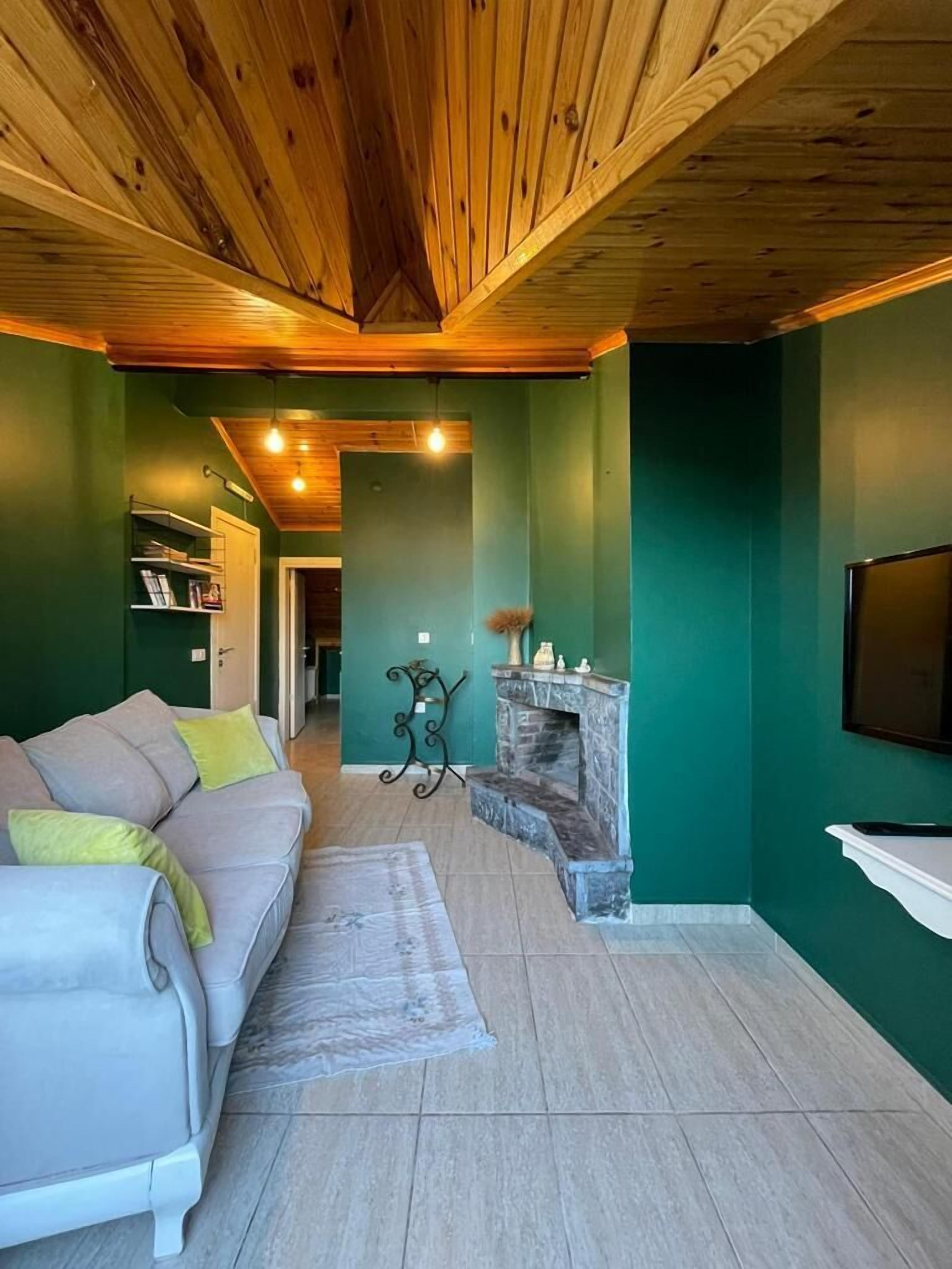 A cozy living room with a wooden ceiling, a green wall, a fireplace, and a sofa with cushions.