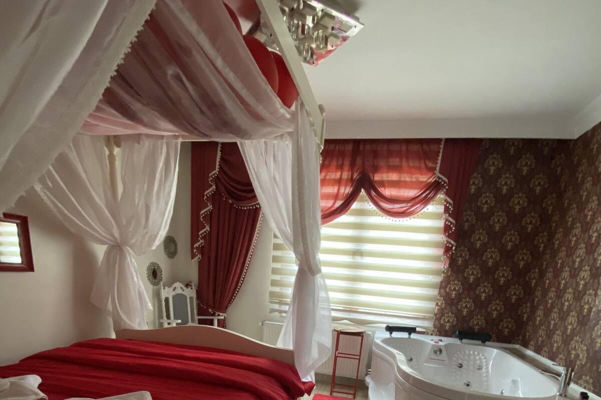 A bedroom with a four-poster bed, a separate sitting area with a chair, a large bathtub, and a window with blinds.