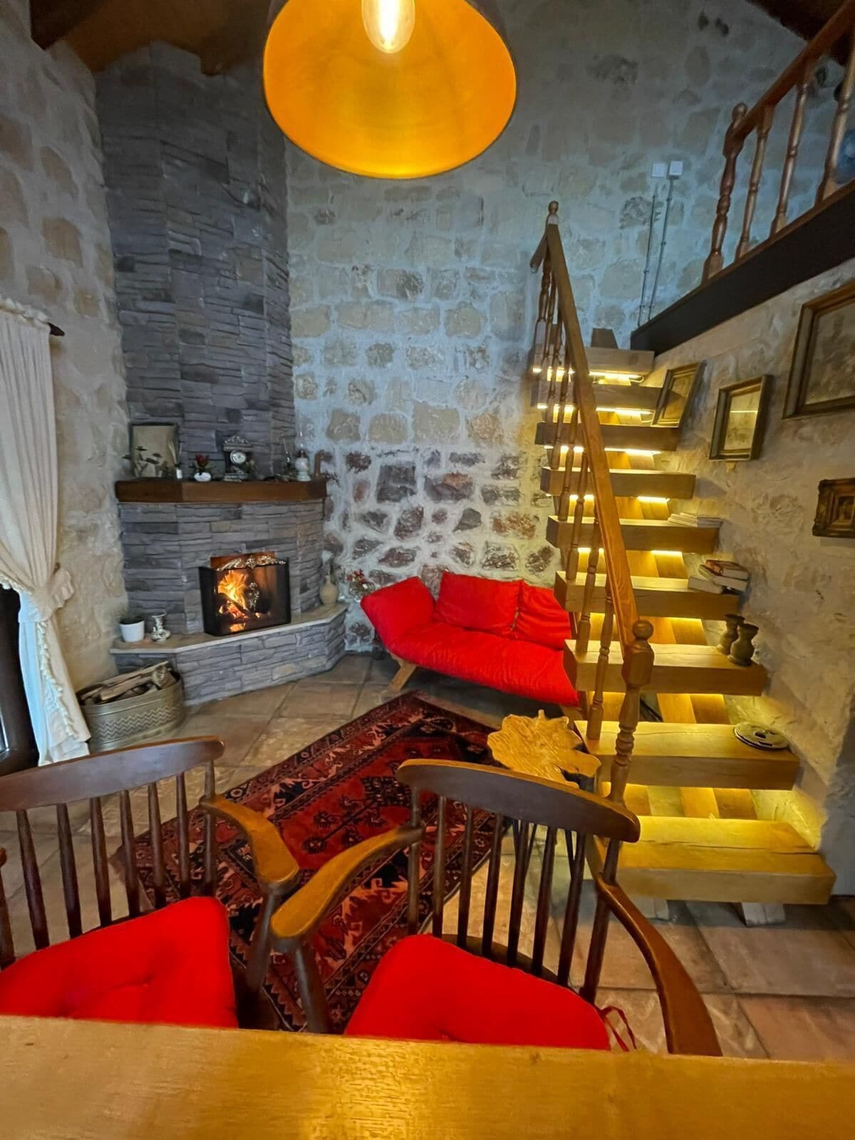 A cozy interior with a stone wall, a fireplace, a wooden staircase, and a red sofa.