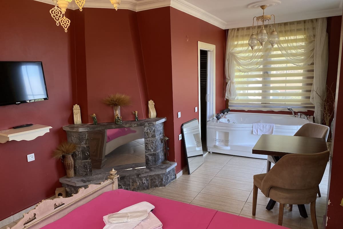 A bedroom with a TV, a fireplace, a bed, a chair, and a bathroom with a bathtub and window.