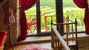 Una habitación con techo de madera, un maniquí, una silla roja y vistas a un paisaje verde a través de amplios ventanales con cortinas rojas.