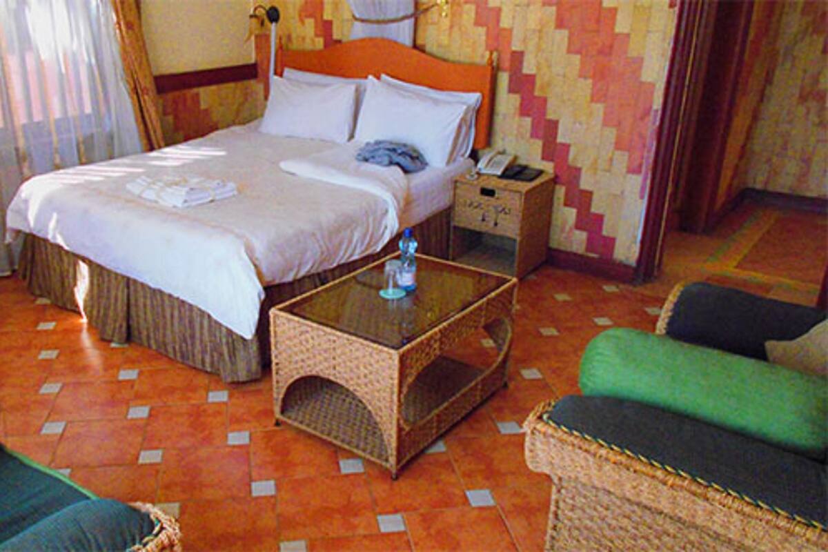 A hotel room with a bed, a bedside table with a bottle, a nightstand, and two green chairs.