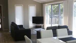 A modern living room with a flat-screen TV, two sofas, a dining table, and large windows. - Malbecs (Lake Tyers Beach)