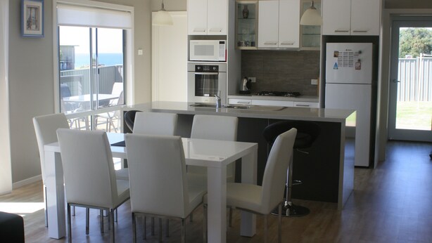 A modern kitchen with white cabinetry, a central island, and a dining area with white chairs. - Malbecs (Lake Tyers Beach)