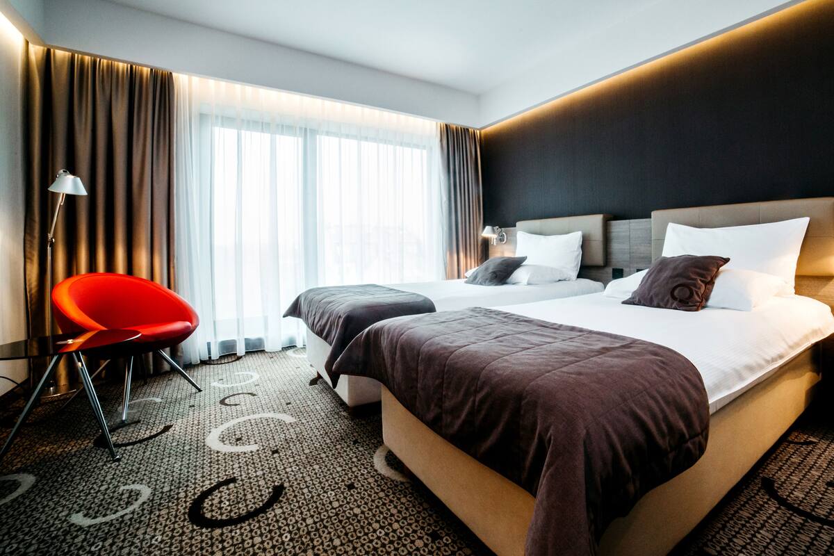 A modern hotel room with a large bed, a red chair, and a large window with curtains.