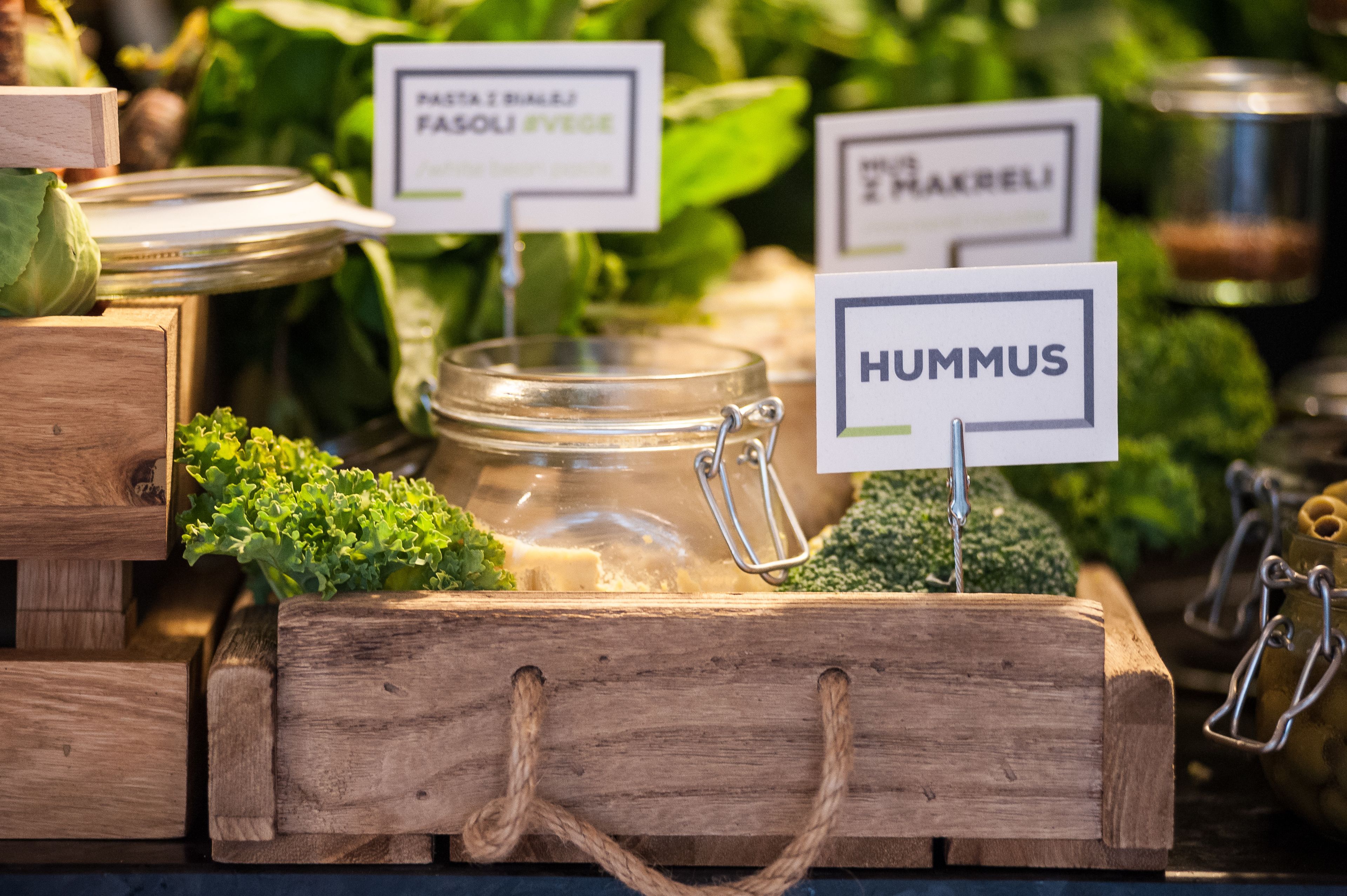 A wooden crate with labeled jars containing various vegetables like hummus, feta, and carrots.