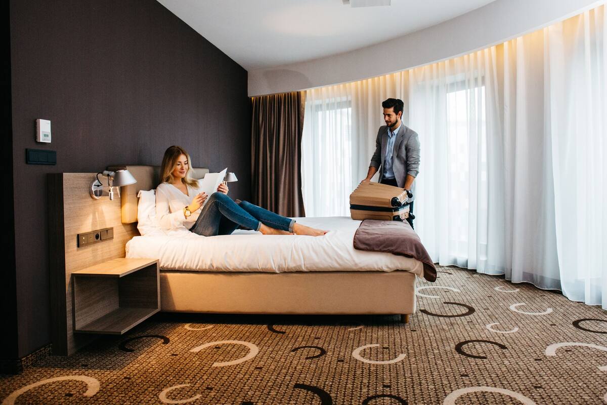 A hotel room with a bed, a bedside table, a lamp, a wall-mounted thermostat, and a large window with curtains.