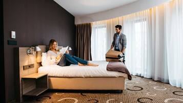 A hotel room with a bed, a bedside table, a lamp, a wall-mounted thermostat, and a large window with curtains.