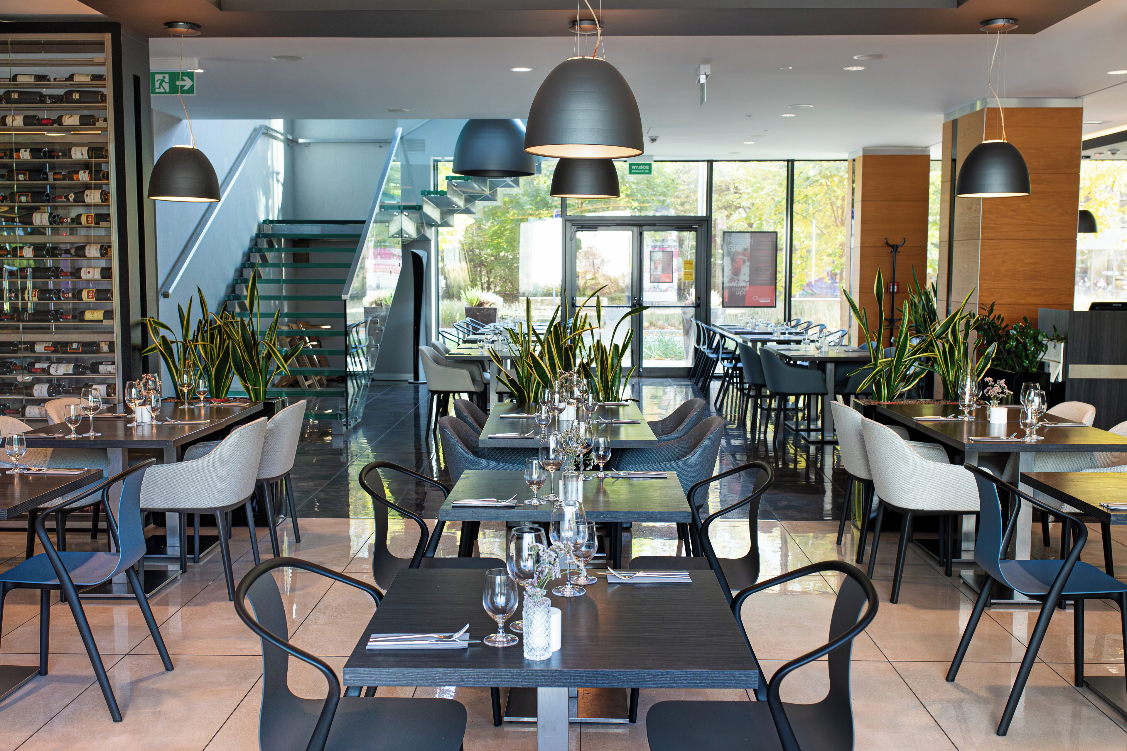 Un restaurante moderno con varias mesas y sillas, una escalera y ventanas amplias.