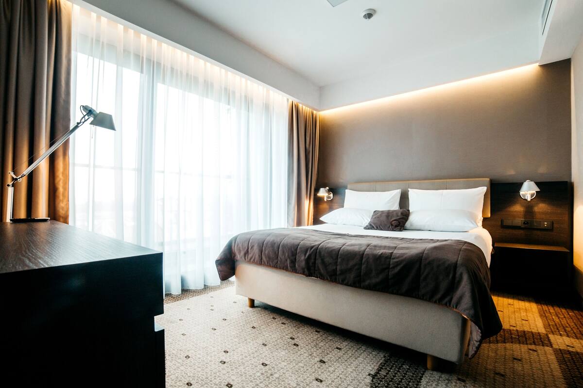 A modern hotel room with a large bed, a desk, a lamp, and a view of the city through sheer curtains.