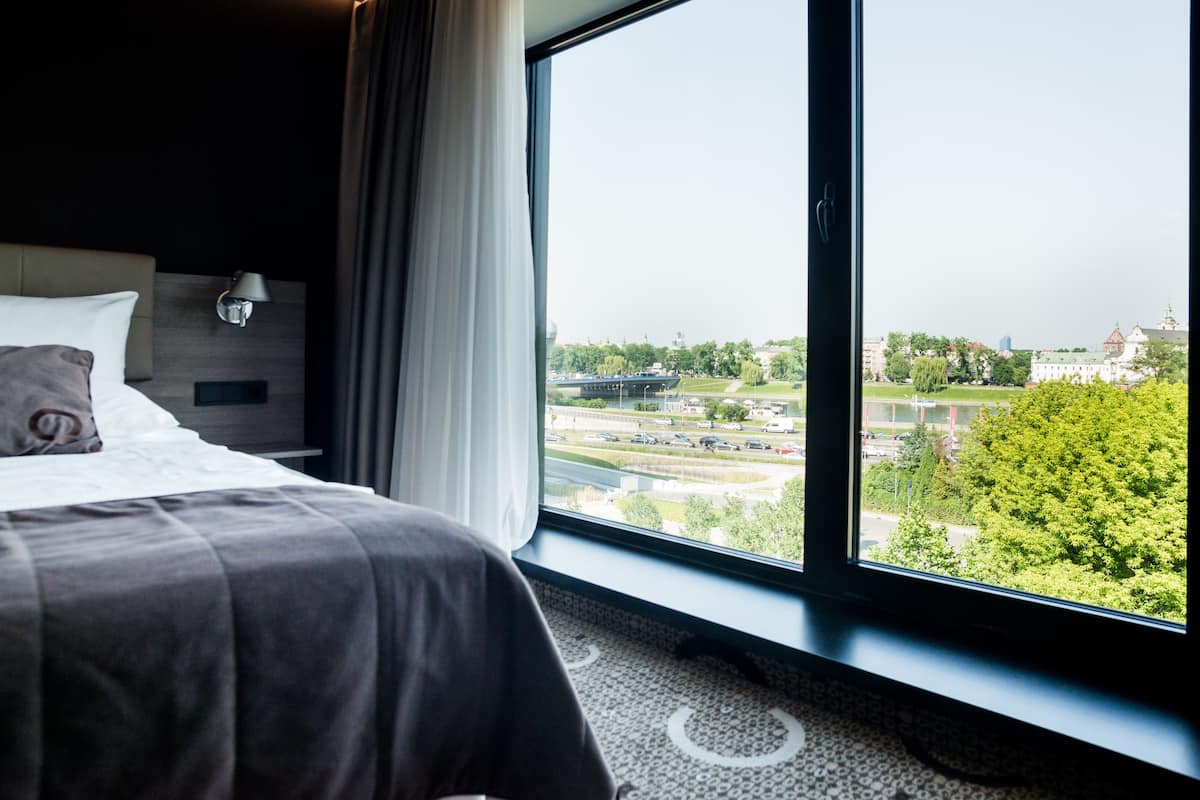 A hotel room with a large window, a bed, a headboard, and a lamp.