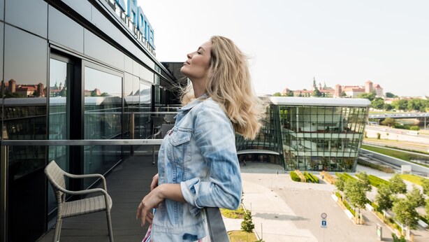 Une femme se tient sur un balcon, d’où l’on peut admirer un bâtiment moderne doté de grandes fenêtres en verre et d’une enseigne indiquant le mot « CENTRE ».