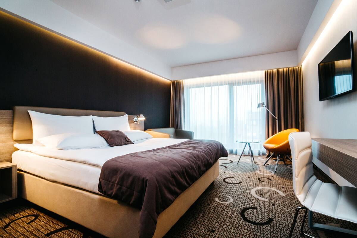 A modern hotel room with a large bed, a desk, a chair, a TV, and a window with curtains.