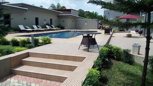 A pool area with lounge chairs and a table, surrounded by greenery and a tiled deck. - Golden Tulip Ibadan (Ibadan)