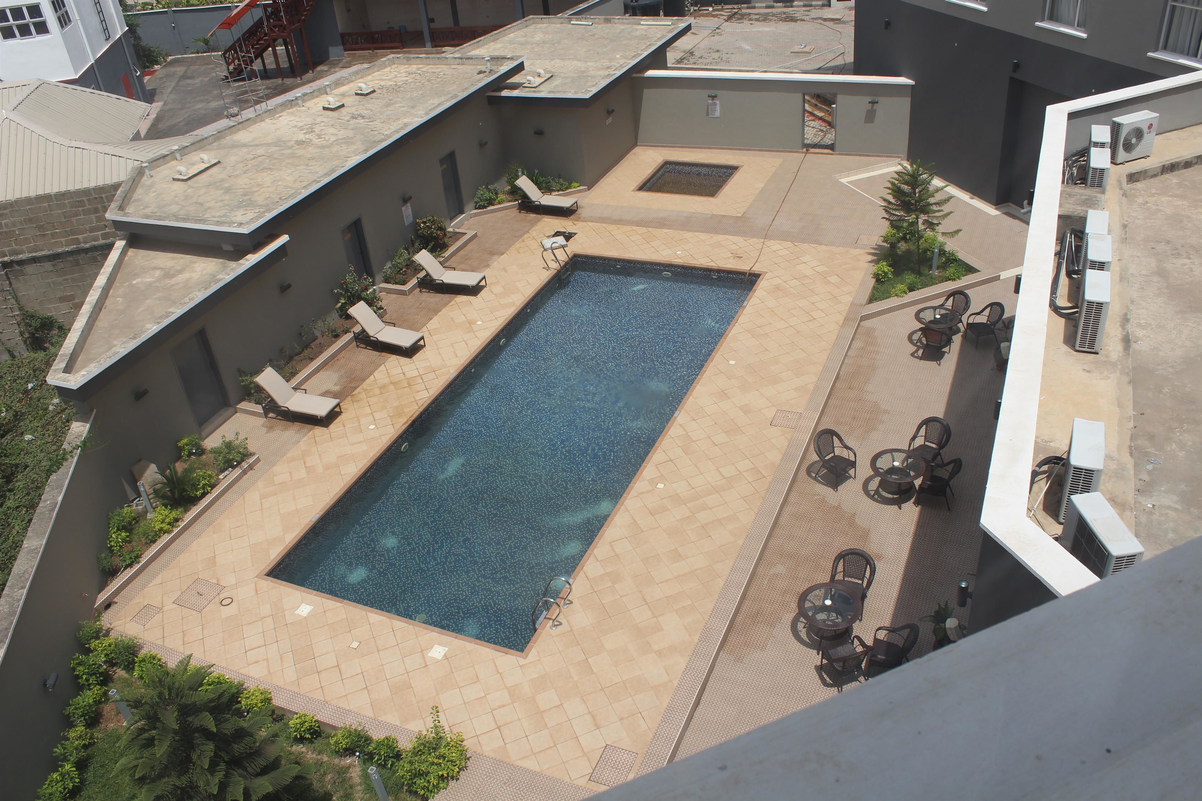 An aerial view of a rooftop swimming pool with lounge chairs and tables, surrounded by buildings.