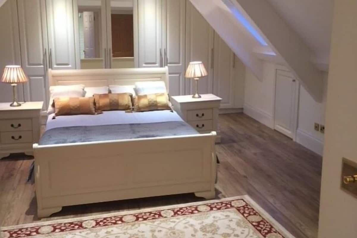 A bedroom with a white bed, two bedside tables with lamps, a mirror above the bed, and a patterned rug on the floor.