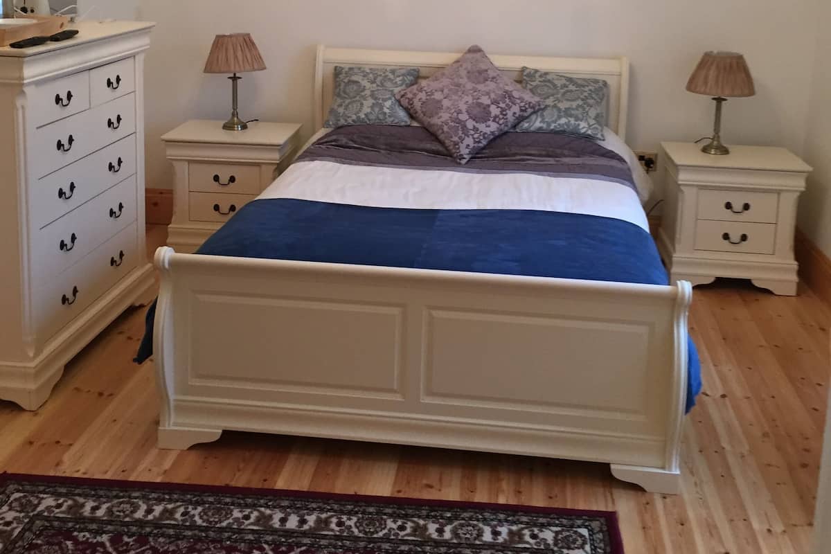 A bedroom with a white four-poster bed, two bedside tables with lamps, a white dresser with multiple drawers, and a patterned rug on the wooden floor.