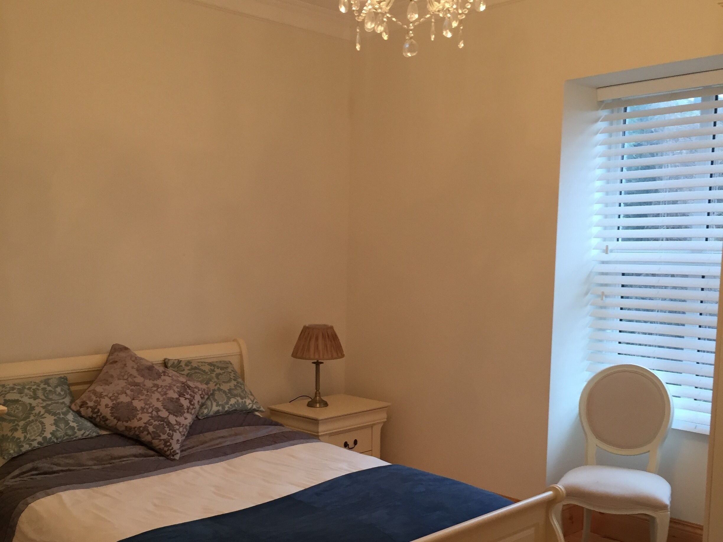A bedroom with a bed, bedside lamp, chair, and window with blinds.
