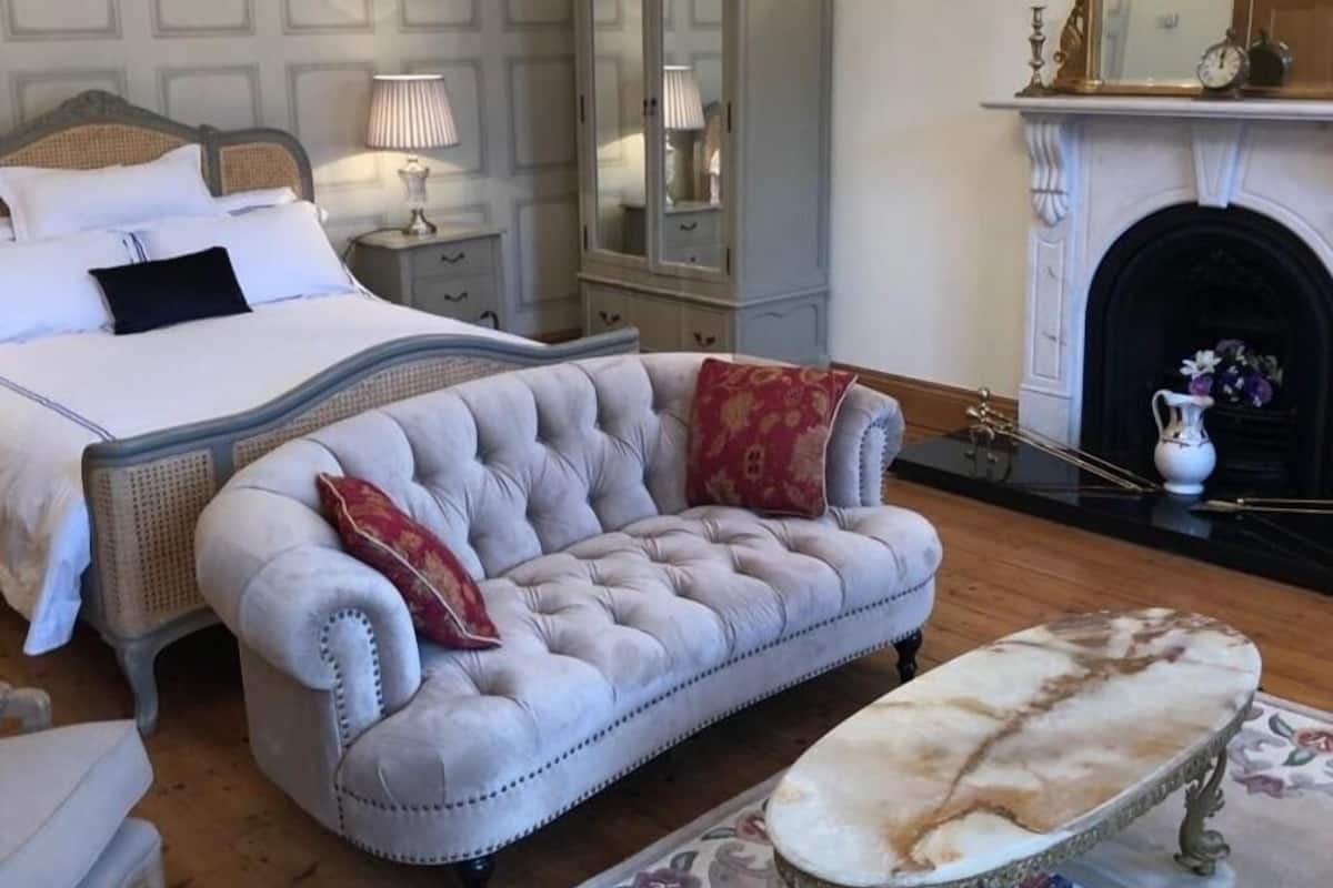 A bedroom with a bed, a chaise lounge, a side table, a mirror, a cabinet, a fireplace, and a chandelier.