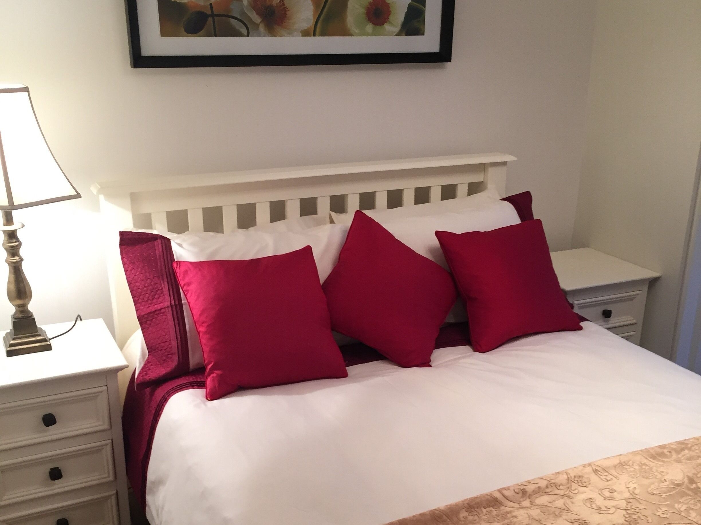 A bed with white bedding and four red pillows, a white nightstand with a lamp, and a framed picture on the wall.