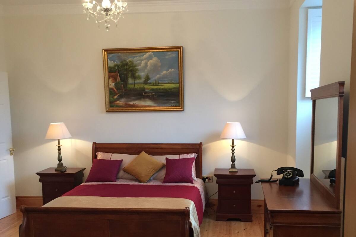 A bedroom with a wooden bed, two bedside tables with lamps, a nightstand with a telephone, a painting on the wall, and a mirror on the right.
