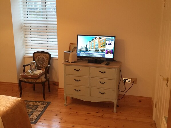 A room with a wooden floor, a white dresser with drawers, a chair, a small table with a tray of items, and a television on top of the dresser displaying a news channel. - The Old Dispensary (Kinsale)