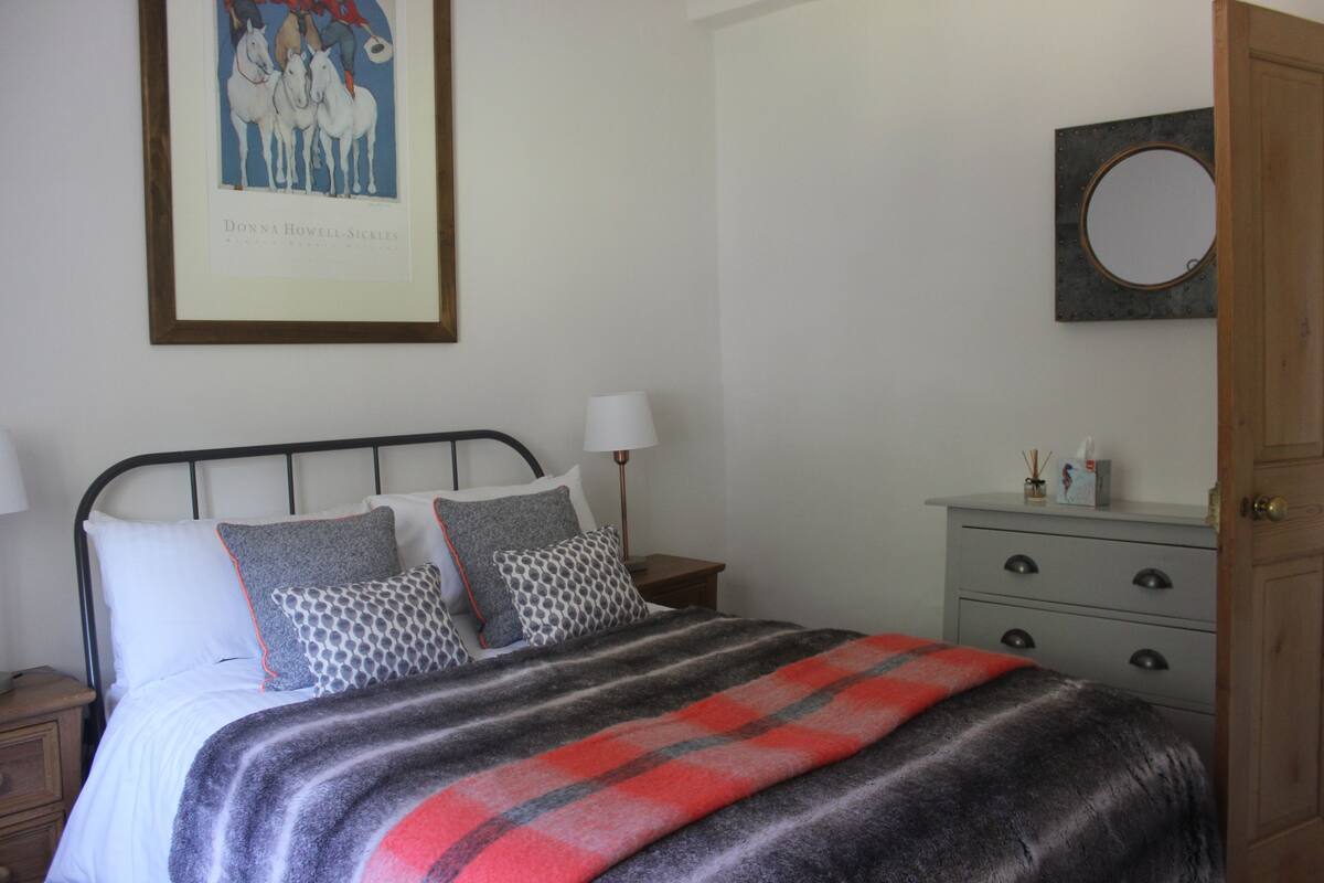 Een netjes opgemaakt bed met een gestreept dekbed, een ingelijst beeld aan de muur, een nachtkastje met een lamp, en een houten deur.
