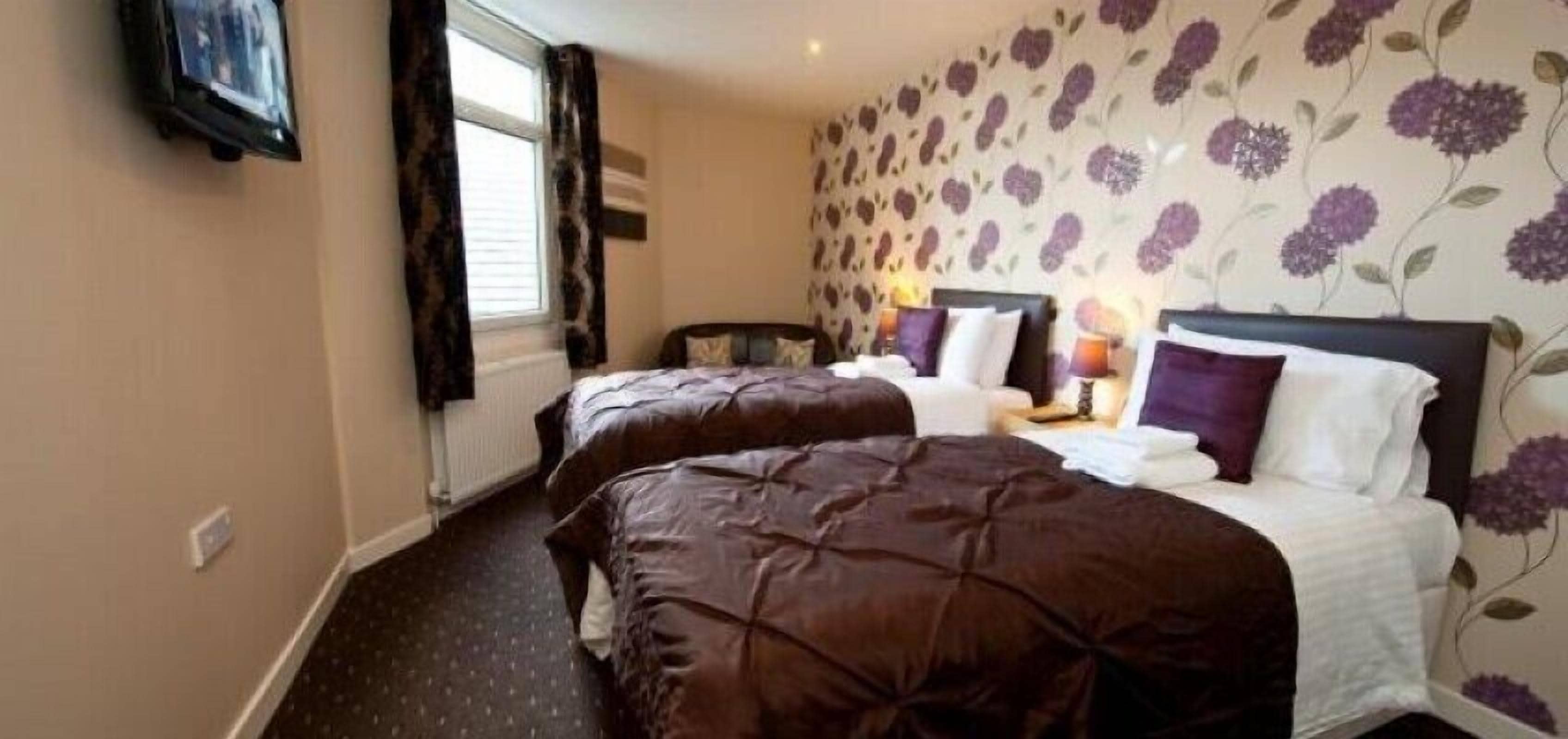 A hotel room with two beds, a nightstand with a lamp, and floral wallpaper.