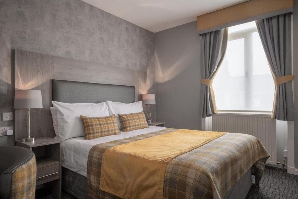A hotel room with a bed, nightstands, lamps, a window with curtains, and a radiator.