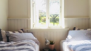 A bedroom with two beds, a window with a view of greenery, a bedside table with a lamp and a plant, and a radiator under the window.