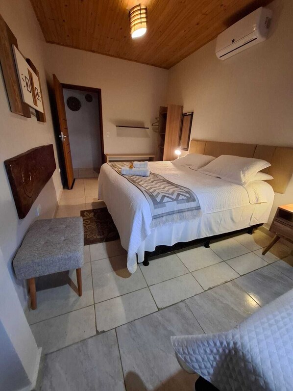 A bedroom with a bed, a bench, a bedside table, a wall-mounted air conditioner, and a door.