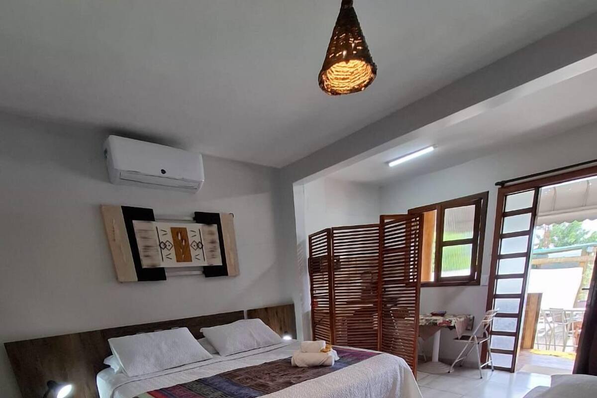 A bedroom with a bed, bedside table, lamp, air conditioning unit, decorative wall art, and a view of the outside.