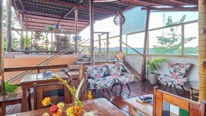 Un espacio de estar al aire libre con techo, mesas y sillas de madera, suelo de baldosas y vistas al mar.