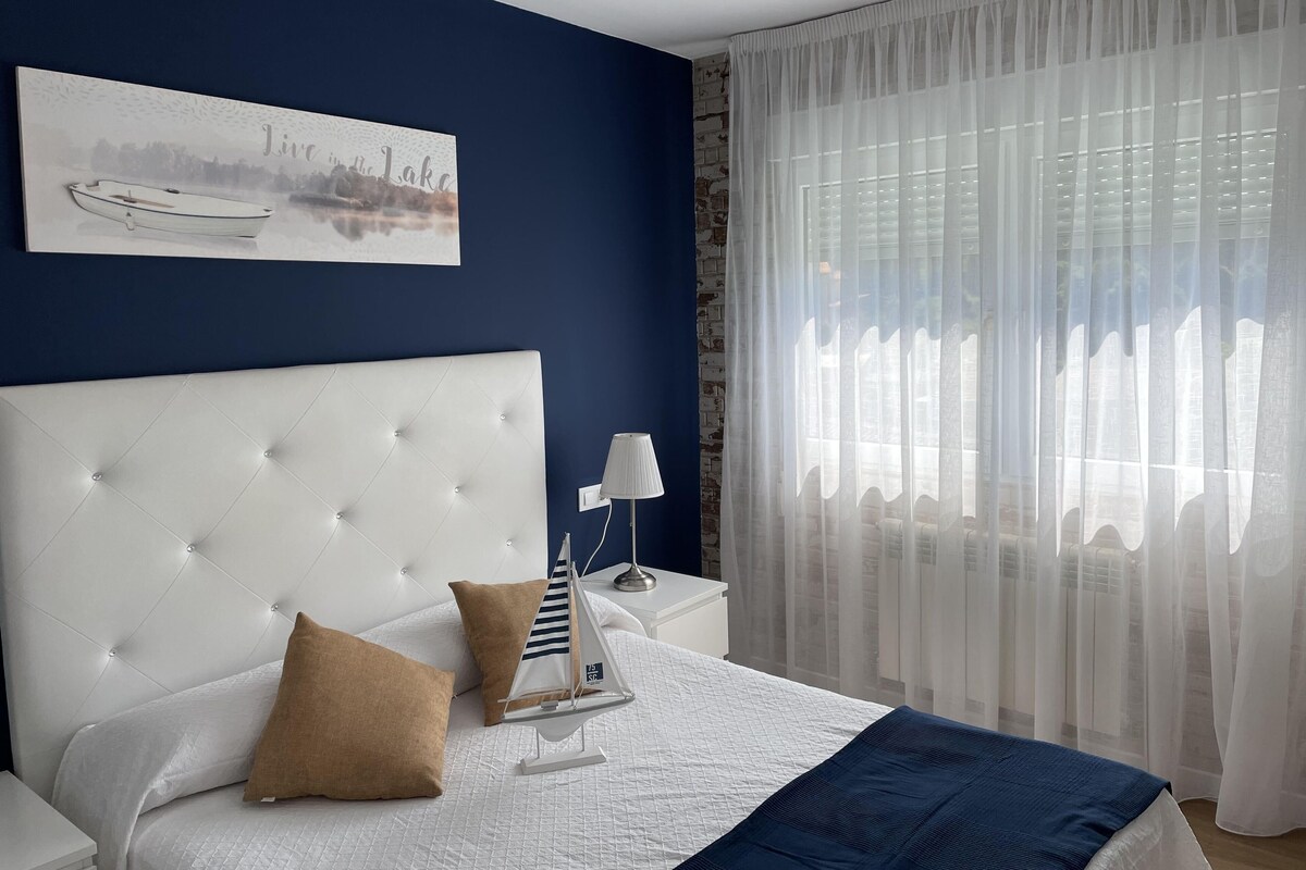 Ein Schlafzimmer mit einem weißen, gepolsterten Kopfende, einem Bett mit weißer und blauer Bettwäsche, einem Modell eines Segelbootes und einem Gemälde einer Seenlandschaft.