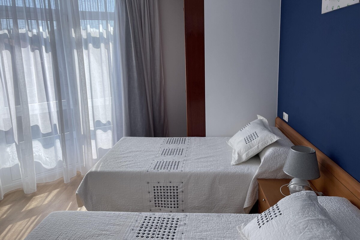 Ein Doppelbett mit Holz-Kopfstück, ein Nachttisch mit Lampe und durchsichtige Vorhänge.
