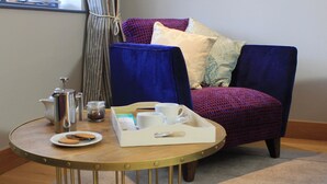 Un tavolo rotondo con un vassoio contenente un teiera, tazze e biscotti, posto di fronte a una poltrona blu con cuscini decorati.